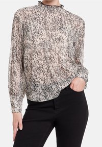Blouse longue transparente à manches longues beige clair avec un motif moucheté noir, col à volants et poignets froncés, associée à un pantalon noir.