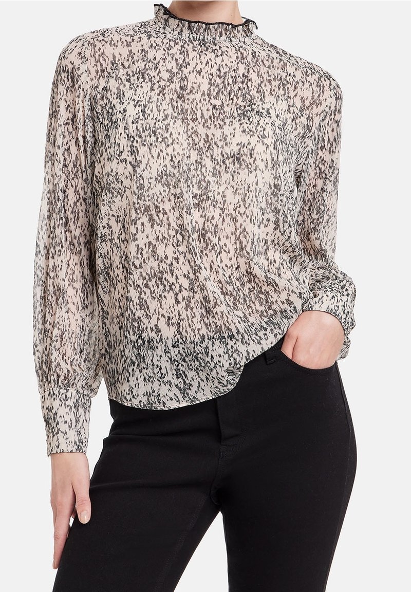 Blouse longue transparente à manches longues beige clair avec un motif moucheté noir, col à volants et poignets froncés, associée à un pantalon noir.