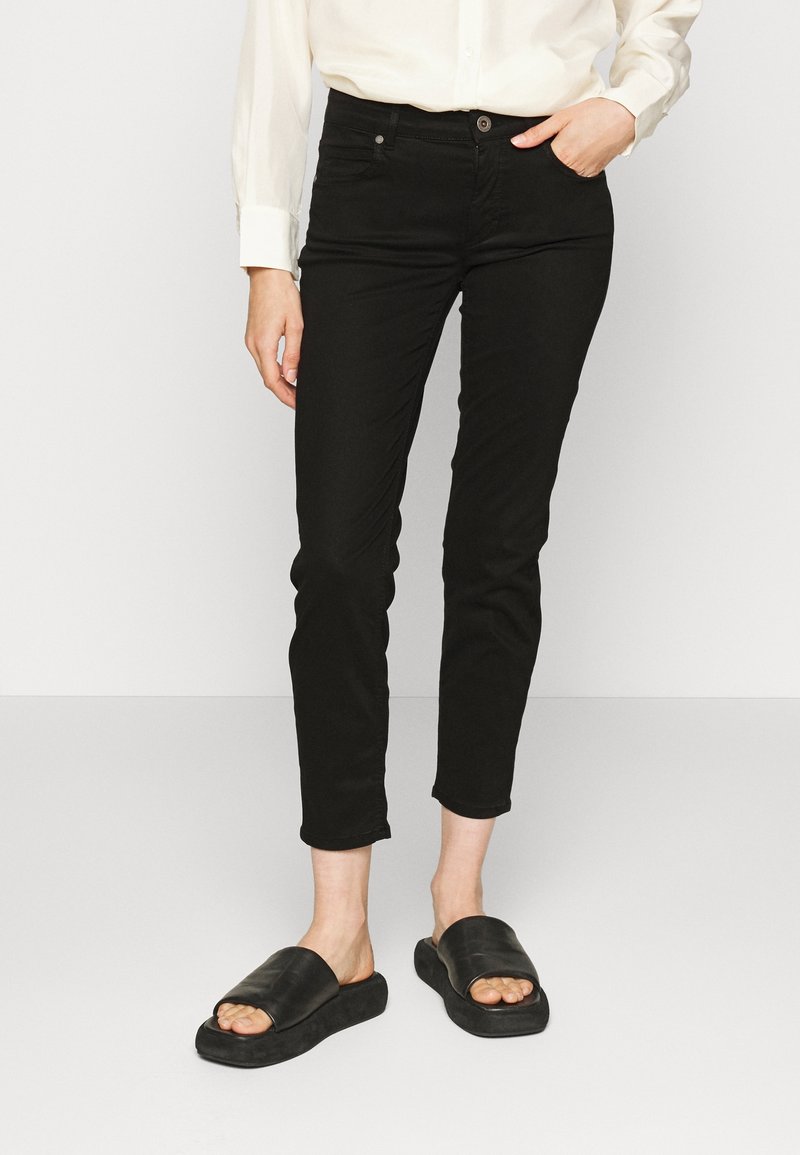 Personne portant un pantalon slim noir, une chemise blanche à manches longues et des sandales noires à enfiler, debout avec une main dans la poche.