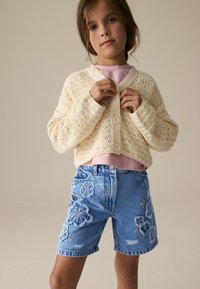 Cardigan en tricot couleur crème avec un col en V et une fermeture à boutons, associé à un short en denim bleu avec des broderies florales et des accents déchirés.