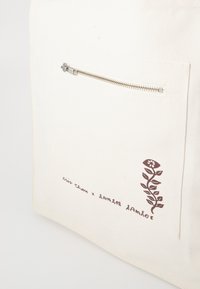 Canvas tote bag in off-white with a silver zipper. Features a dark brown floral design and text near the bottom left corner.
