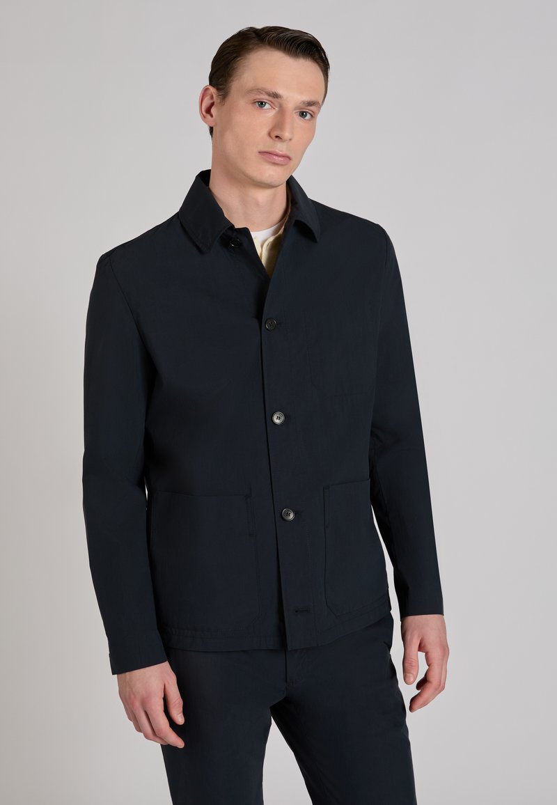 Jeune homme portant une veste noire boutonnée et un pantalon assorti, debout devant un fond gris clair uni.