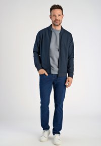 Granatowa kurtka bombowiec z stójką, szary sweter pod spodem, niebieskie dżinsy i białe trampki. Gładki materiał o minimalistycznym designie.