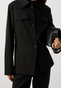Czarna kurtka z zapięciem na guziki z przodu, dwoma dużymi kieszeniami na piersi i kołnierzem, zestawiona z czarną, teksturowaną kopertówką.