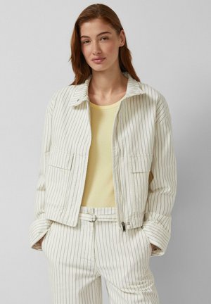 Frau trägt eine cremefarbene Nadelstreifenjacke und passende Hose über einem blassgelben Oberteil, steht mit den Händen in den Taschen vor einem einfarbigen Hintergrund.
