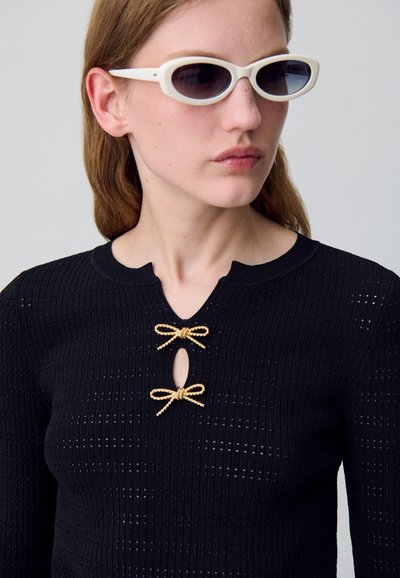 Top negro de punto con un escote en forma de ondas, que cuenta con dos lazos dorados y un corte ovalado. La modelo lleva gafas de sol blancas.