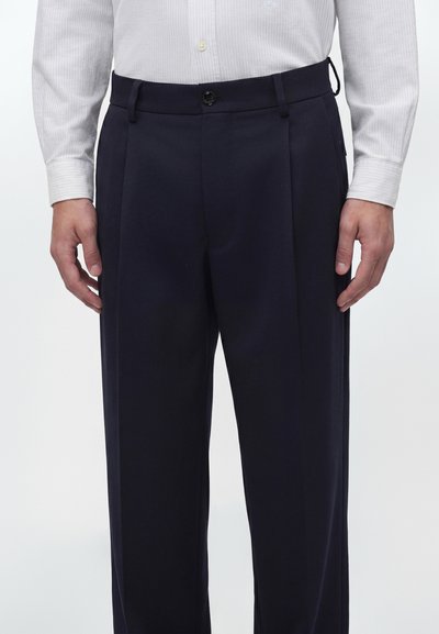 Homme portant un pantalon de costume plissé bleu marine foncé avec un bouton noir et une chemise blanche à manches longues rentrée, debout, les mains détendues sur les côtés.