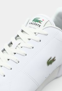Sapatilha de pele branca com atacadores brancos planos, costura decorativa e um logo de crocodilo verde da Lacoste de lado.