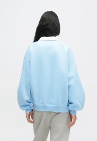 Osoba s dlhými čiernymi vlasmi, oblečená v svetlo modrom oversized mikinovom svetre a šedých nohaviciach, stojí chrbtom k jednému farebnému bielemu pozadiu.