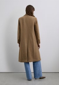Brown wool coat with a straight cut, long sleeves, and three-button cuffs, paired with light blue flared jeans and pointed brown shoes.