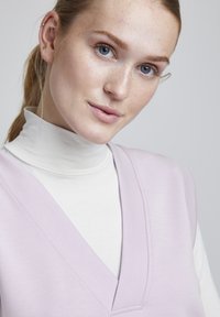Top sin mangas de color púrpura claro con un escote en V profundo, superpuesto a un suéter de cuello alto blanco. Textura suave, acentos mínimos y un diseño limpio.