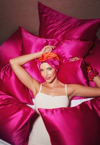 Cojines de satén en un vibrante color rosa apilados, con un modelo que lleva un pañuelo de satén naranja y rosa, mostrando texturas suaves y colores contrastantes.