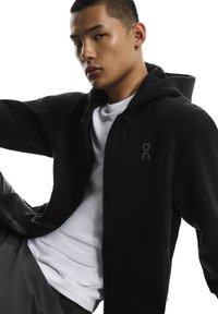 Musta vetoketjullinen huppari, joka on valmistettu pehmeästä ja joustavasta kankaasta. Edessä on vetoketju, säädettävä nyörillinen huppu sekä rintakehällä logo.