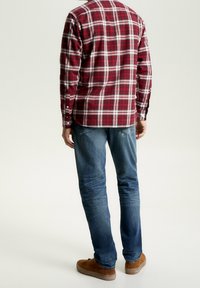 Un homme portant une chemise à carreaux rouge et blanc, un jean bleu et des chaussures marron se tenant de dos face à l'appareil photo, sur un fond uni.