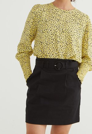 Femme portant un chemisier jaune à manches longues avec des taches noires, rentrée dans une jupe noire ceinturée, les mains dans les poches.