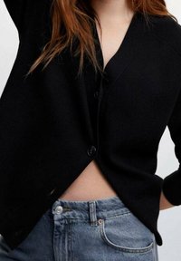 Černý cardigan s výstřihem do V, zapínáním na knoflíky vzadu a mírně oversized střihem. V doplnění s modrými džíny s vysokým pasem.