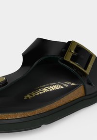 Μαύρο σανδάλι Birkenstock με φελλό σόλα και ρυθμιζόμενο χρυσό αγκράφα σε λευκό φόντο.