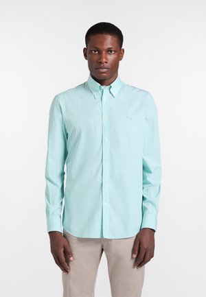 Mann trägt hellblau gestreiftes Hemd mit Knöpfen und beige Hose, steht frontal vor weißem Hintergrund.