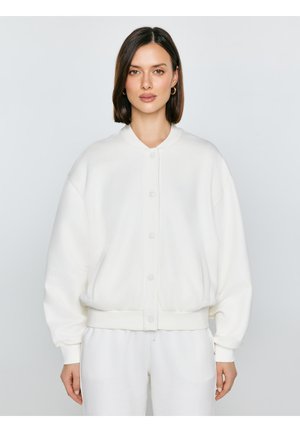 Kobieta z prostymi brązowymi włosami nosi białą bomberkę zapinaną na guziki i białe spodnie, stoi na tle jednolitego jasnoszarego tła.