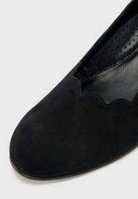 Black suede flat shoe with a scalloped edge, rounded toe, and perforated inner lining, featuring a smooth, matte texture.