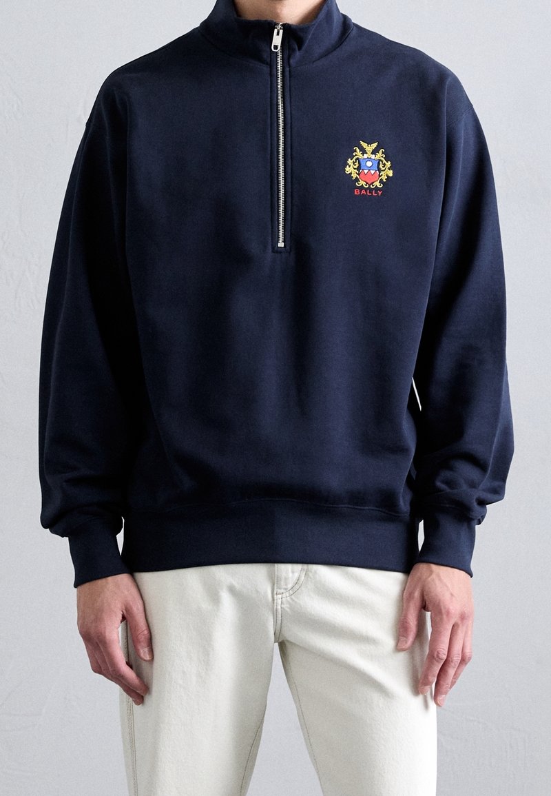 Man wearing navy blue half-zip sweatshirt with colorful crest and red "BALLY" text, paired with off-white pants.