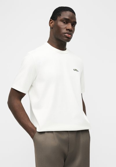 Άνδρας που φοράει λευκό κοντομάνικο πουκάμισο με μικρό λογότυπο στο στήθος και καφέ παντελόνι, στέκεται μπροστά σε απλό ανοιχτό γκρι φόντο με τα χέρια στις τσέπες.