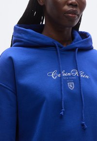 Modrá mikina s kapucňou z bavlneného zmesi s nastaviteľnou šnúrkou. Na hrudi je biely vyšívaný nápis "Calvin Klein" a motív štítu.
