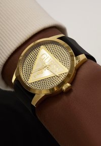 Montre dorée avec un cadran triangulaire présentant une texture en maille, un bracelet en silicone noir et un logo Guess visible avec un détail de point d'interrogation.