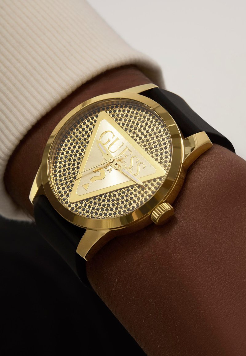Montre dorée avec un cadran triangulaire présentant une texture en maille, un bracelet en silicone noir et un logo Guess visible avec un détail de point d'interrogation.