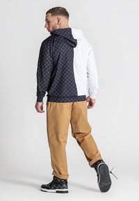 Hooded dukserica s odvojenim dizajnom: na jednoj strani crna šarena tkanina, a na drugoj strani bijela šarena tkanina. U paru s bež hlačama.