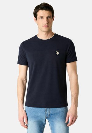 Uomo che indossa una maglietta blu navy semplice con un piccolo logo ricamato sul petto e jeans azzurri, in piedi davanti a uno sfondo bianco.