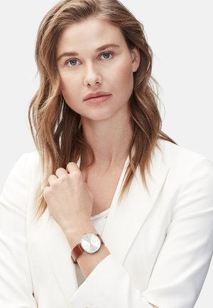 Brauner Lederarmbanduhr mit einem runden silbernen Zifferblatt, goldenen Akzenten und einer glatten Oberfläche, getragen an einem Handgelenk mit einem weißen Blazer.