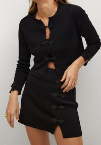 Mustavillinen neulecardigan, jossa on pyöreä pääntie, nilkkapituiset hihat ja koristeelliset hakasulkimet. Täydentävässä hameessa on sivureikä ja samanlaiset sulkimet.