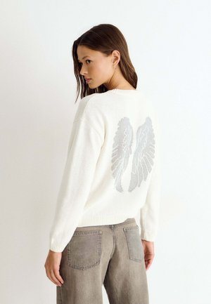 Mujer con suéter blanco con diseño de alas de ángel plateadas en la espalda, combinada con jeans grises, de pie frente a un fondo blanco liso.