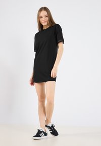 Czarna bawełniana sukienka t-shirty z krótkimi rękawami, ozdobiona koronkowym wykończeniem, w zestawieniu z czarnymi zamszowymi sneakersami z białymi akcentami, na neutralnym tle.