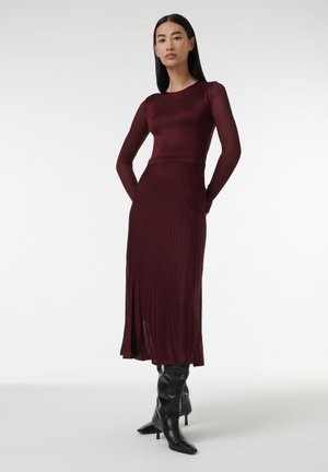 Robe en maille côtelée bordeaux avec des manches longues, un corsage ajusté et une jupe évasée ; associée à des bottes noires à talons hauts et au genou.