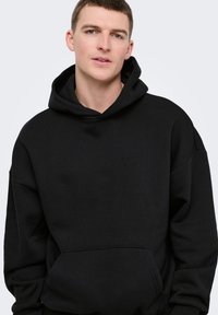 Jeune homme aux cheveux courts portant un sweat à capuche noir uni, sur un fond gris clair, légèrement tourné vers la gauche.