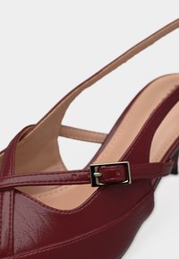 Chaussure slingback en cuir verni bordeaux avec une bride étroite et une petite boucle dorée sur une semelle beige et un talon bas.
