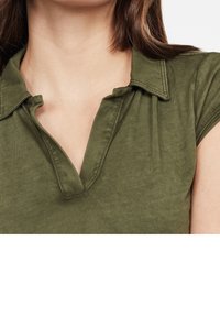 Olivgrön polo med krage och V-ringning, korta ärmar och ett mjukt, strukturerat tyg. Synliga sömdekorationer.