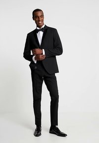 Smoking noir avec un nœud papillon, comprenant une veste sur mesure, un pantalon ajusté et des chaussures de ville noires brillantes, sur un fond neutre.