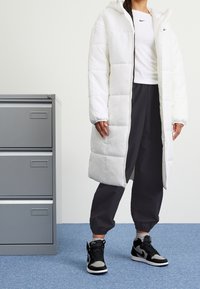 Vit, lång vadderad jacka med huva och quiltad design. Kombinerad med svarta joggers och höga sneakers. Neutrala bakgrundselement.