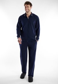 Navy trainingspak van zacht materiaal, bestaande uit een ritsjack met een gekleurd kraag en bijpassende broek met zakken, gedragen met grijze slippers.