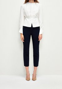 Witte tekstuurbloes met geplooide achterkant, lange mouwen en manchetten. Gecombineerd met marineblauwe broek tot de enkels en naaldhakken in nude.