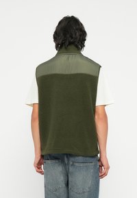 Groene fleece gilet met een gladde bovenkant en een gestructureerde onderkant. Heeft een hoge kraag en een relaxte pasvorm. Spijkerbroek zichtbaar.