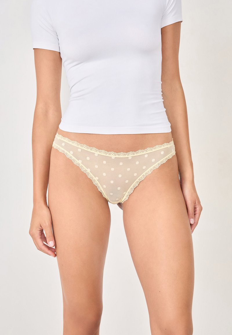 Tanga beige trasparente con pois bianchi, accentuato da una delicata rifinitura in pizzo lungo la cintura e le aperture delle gambe. Texture morbida su tutta la superficie.