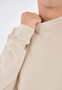Beige langermet sweatshirt med ribbede mansjetter og krage, med en glatt tekstur. Hånd som lett griper om krageområdet.