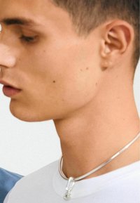 Silverhalsband med ett lås, bärs över en vit T-shirt. Halsbandet har en slät, glänsande yta och en enkel, minimalistisk design.