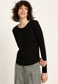 Eine Frau mit kurzen, gewellten braunen Haaren trägt ein schwarzes Langarmoberteil und graue karierte Hosen und posiert vor einem schlichten hellen Hintergrund.