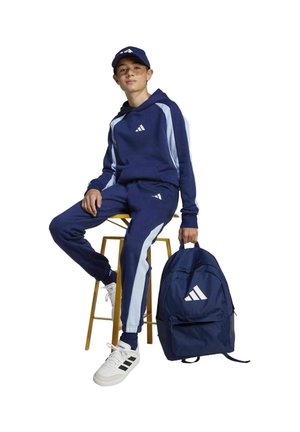 Un joven está sentado en un taburete con un chándal azul marino, una gorra y zapatillas blancas; a su lado hay una mochila a juego de color azul marino.