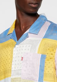 Multikleurige T-shirt met korte mouwen met een mix van paisley- en geometrische patronen in blauw, geel en roze. Beschikt over een borstzak en knoopsluiting.